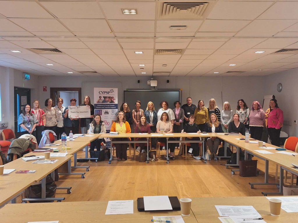 Group photo of approximately 25–30 adults gathered in a meeting room, standing and seated around U-shaped tables. Papers, notebooks, and takeaway coffee cups are placed on the tables. At the centre, two people hold a sign reading “Thank You, Volunteers.” A banner behind them reads “CYPSP – Children & Young People, Parents and Teacher Support.” The room has a wooden floor, white ceiling tiles with lights and vents, and a projector screen at the back.
