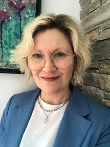 Head-and-shoulders portrait of a person with short light-coloured hair and round glasses, wearing a blue blazer over a white top and a silver necklace. The photo is taken indoors against a stone wall, with a colourful abstract painting visible in the background.