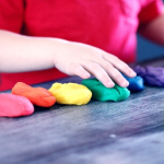 Close-up of a child engaged in a play activity, arranging brightly coloured modelling dough in a row on a table. Red, orange, yellow, green, blue and purple dough pieces are visible, alongside a stack of yellow plastic cups.