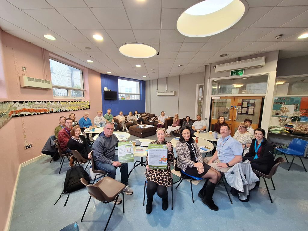 A group of approximately 20 adults seated around tables in a bright community meeting room during a mental health awareness event. Several participants at the front of the room are holding green mental health awareness posters, while others sit around tables with information leaflets and refreshments. The room features pastel pink and blue walls, large windows, circular ceiling lights, and a mosaic artwork displayed along one wall. The photograph captures a collaborative group discussion and community engagement setting focused on promoting mental health awareness and wellbeing.