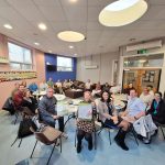 A group of approximately 20 adults seated around tables in a bright community meeting room during a mental health awareness event. Several participants at the front of the room are holding green mental health awareness posters, while others sit around tables with information leaflets and refreshments. The room features pastel pink and blue walls, large windows, circular ceiling lights, and a mosaic artwork displayed along one wall. The photograph captures a collaborative group discussion and community engagement setting focused on promoting mental health awareness and wellbeing.