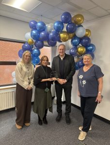 Four members of staff stand together for a presentation photograph beneath a decorative balloon arch in shades of blue, light blue, white and gold. Two people in the centre are holding a glass trophy awarded for Partnership and Driving Success. The photograph was taken in an office environment during the SPPG Staff Awards and Recognition Event 2026 and captures colleagues celebrating the Children’s Services Planning Team’s award-winning work to strengthen partnership working and improve outcomes for children, young people and families across Northern Ireland.