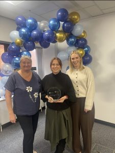 Three members of the Children’s Services Planning Team pose with an award at the SPPG Staff Awards and Recognition Event 2026. The person in the centre is holding a glass trophy for the Partnership and Driving Success Award, while standing beneath a decorative arch of blue, light blue, white and gold balloons. The photograph was taken indoors in an office setting and celebrates the team’s achievement in partnership working and service improvement.
